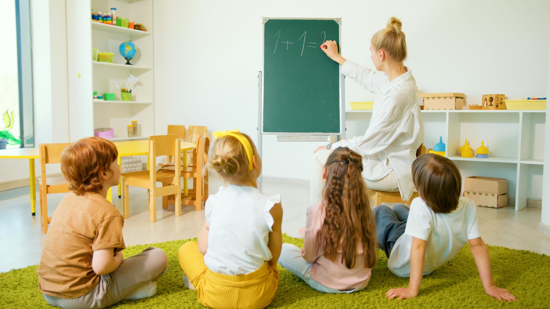 Teacher instructing young students with math on a chalkboard in a bright classroom setting.